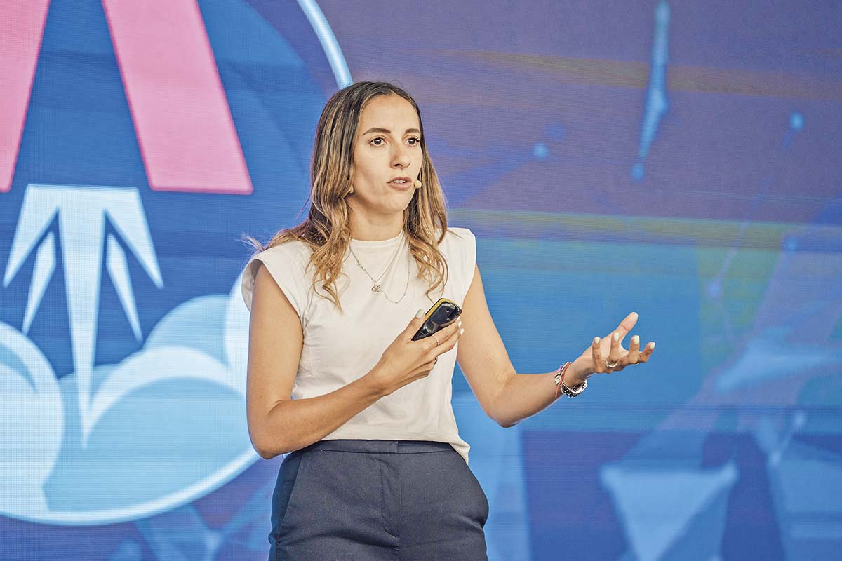 Mujer presentando en una conferencia, de pie frente a una audiencia, con una pantalla de proyección detrás.
